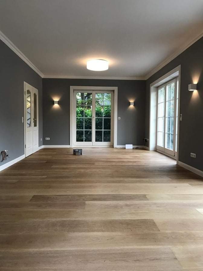 Wnętrze pokoju z nowo ułożoną, jasną drewnianą podłogą, szarymi ścianami, białymi listwami przypodłogowymi i oknami balkonowymi wychodzącymi na zieleń.