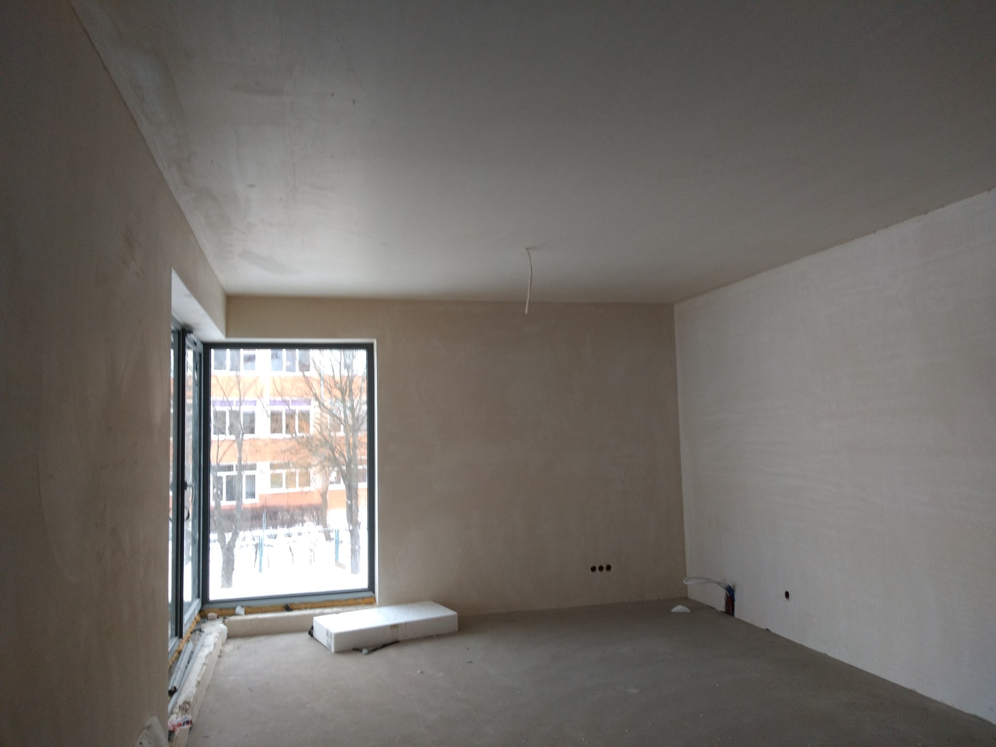 Surowe wnętrze pokoju w trakcie prac wykończeniowych, ściany po szpachlowaniu, widoczne gniazdka elektryczne i okno z widokiem na zimowy krajobraz.