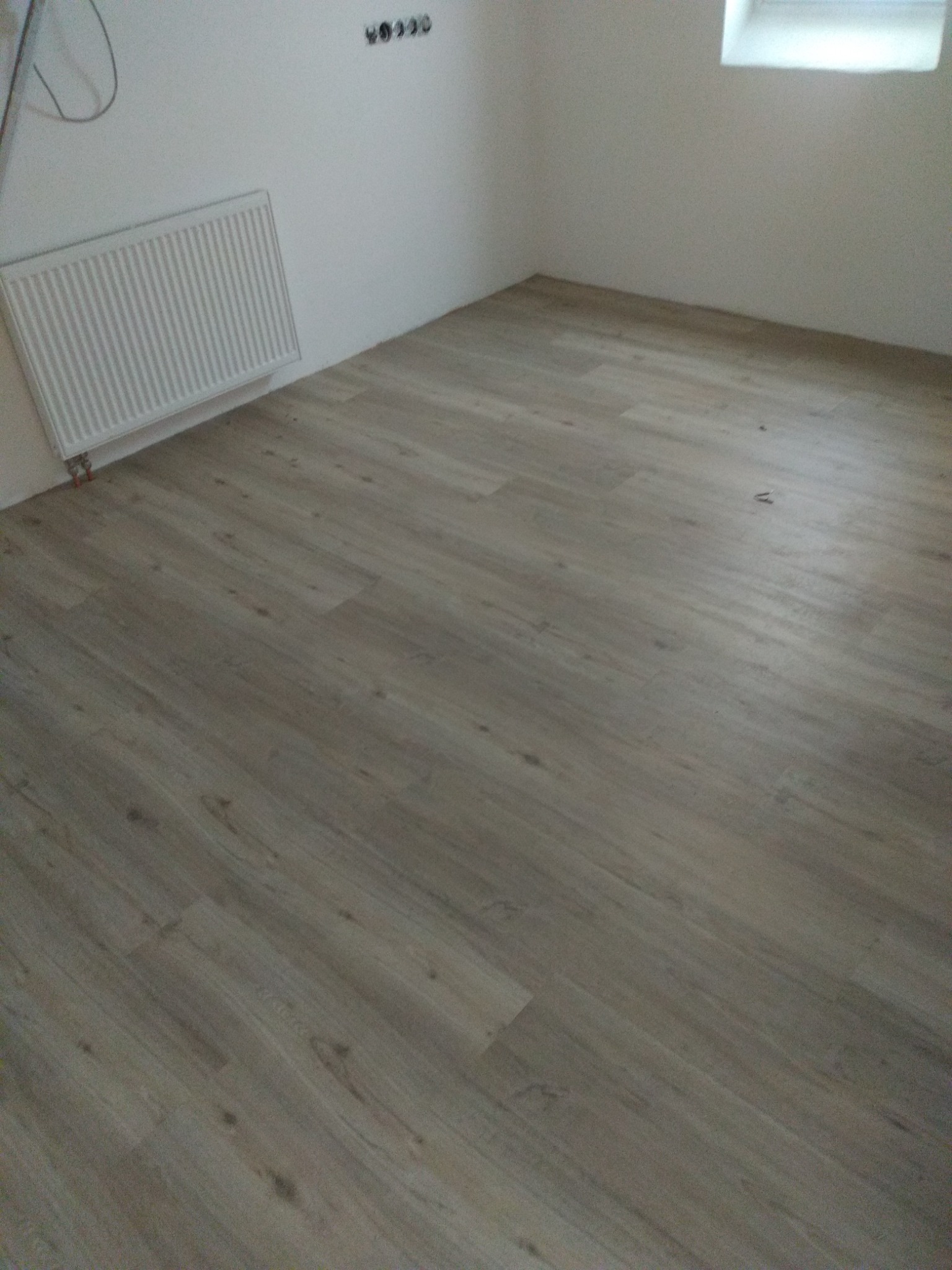Pokój po remoncie z panelami podłogowymi w odcieniach szarości i beżu, białe ściany z grzejnikiem i gniazdkami elektrycznymi, małe okno w górnym rogu.