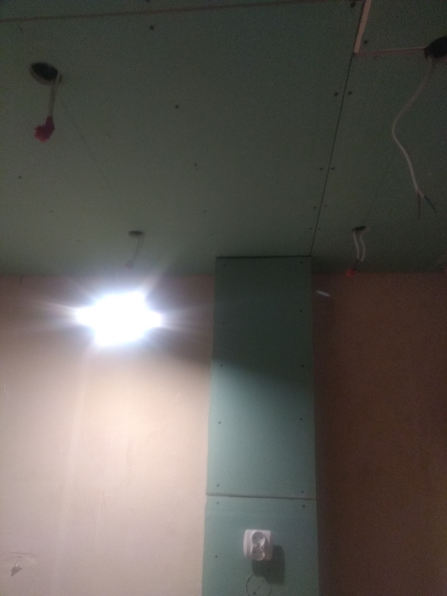 Montaż zielonej płyty gipsowo-kartonowej na suficie i ścianie, widoczne kable elektryczne, gniazdko i oświetlenie robocze.