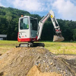 Mała koparka Takeuchi TB216, biało-czerwona, stoi na hałdzie ziemi i gruzu na tle zielonego wzgórza i błękitnego nieba z chmurami.