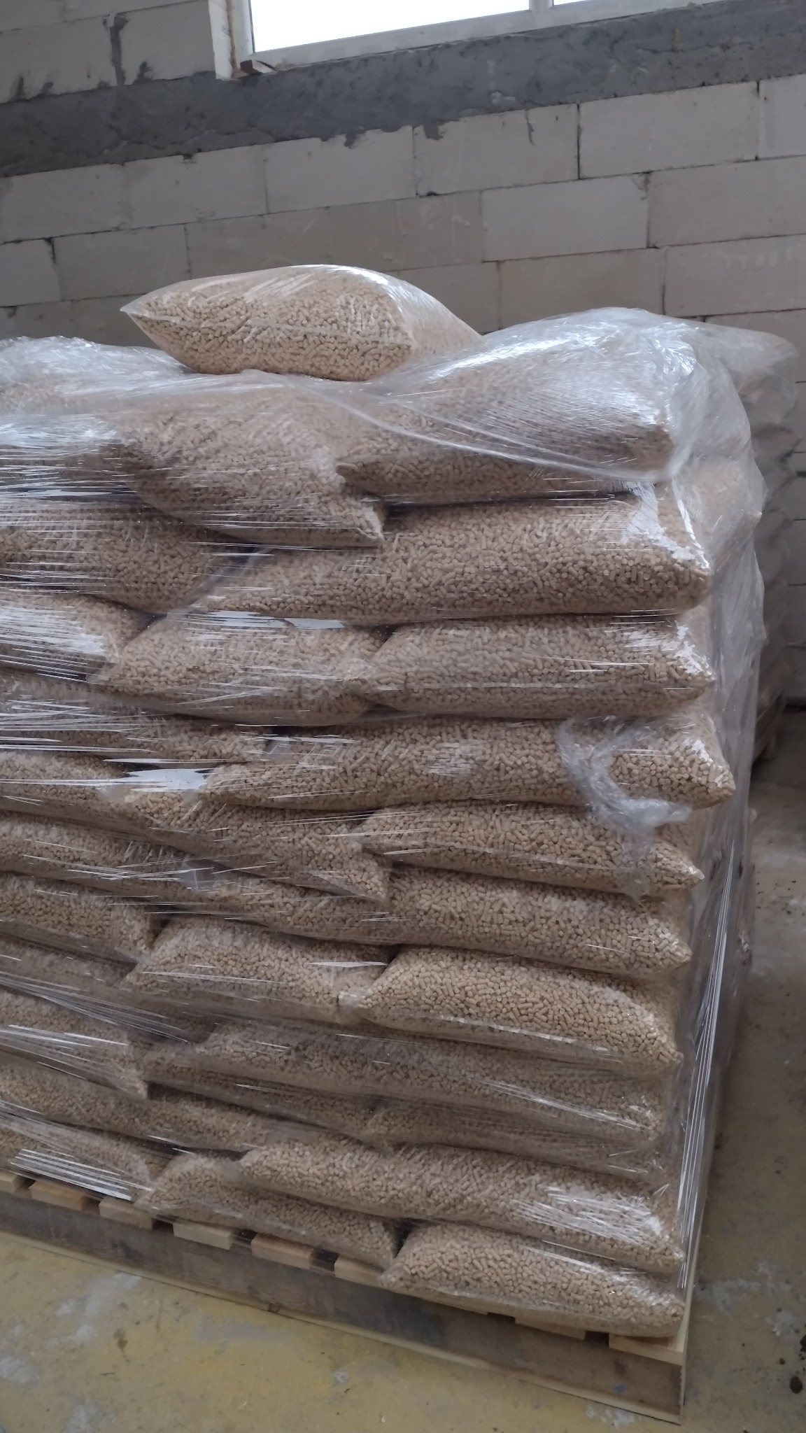 Stos worków z pelletem drzewnym, owiniętych folią stretch, ułożonych na palecie w magazynie z betonowymi ścianami.