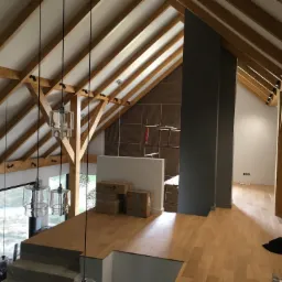 Wnętrze poddasza z drewnianą konstrukcją więźby dachowej, oświetleniem punktowym i wiszącymi lampami, widok z antresoli z drewnianą podłogą i kartonami.