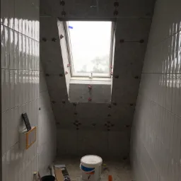 Montaż płytek na ścianach i skośnym suficie łazienki z oknem dachowym, widoczne poziomice laserowe i plastikowe krzyżyki dystansowe, na podłodze resztki kleju i puste wiadra.