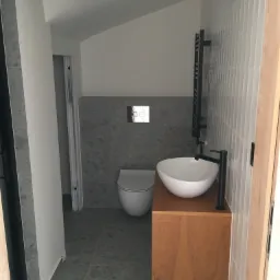 Nowoczesna łazienka z umywalką nablatową na drewnianej szafce, szarą mozaiką na ścianie za toaletą i grzejnikiem drabinkowym w kolorze antracytowym.