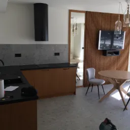 Nowoczesna kuchnia z drewnianymi szafkami, czarnym blatem i okapem, szarą mozaikową ścianą nad blatem, oraz stołem z krzesłami na szarej podłodze z mozaiki, widok na salon przez drewniane drzwi...