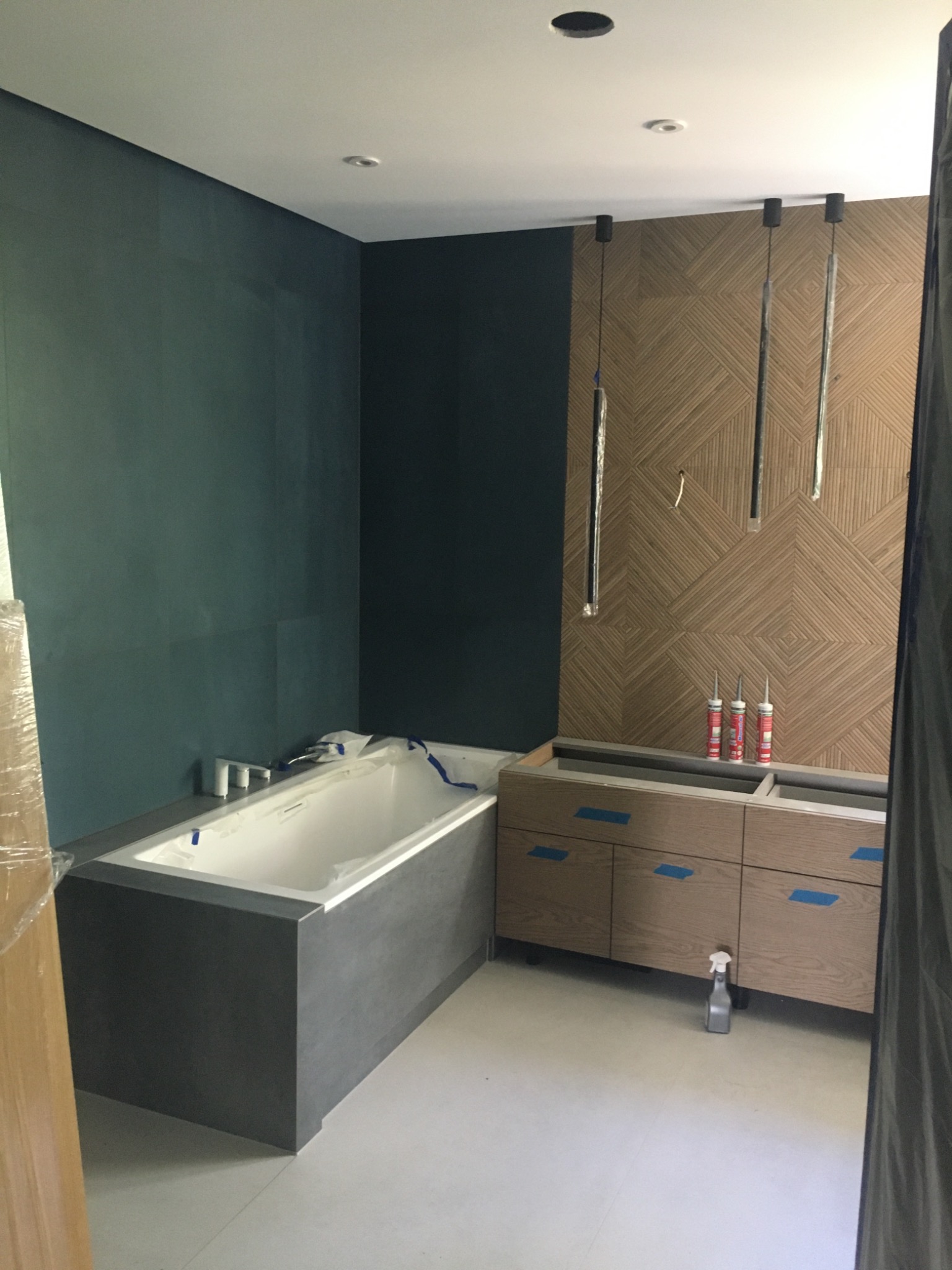 Łazienka w trakcie remontu: zabudowana wanna z szarą obudową, szafka pod umywalkę z okleiną drewnopodobną zabezpieczona taśmą, ściana z płytek o geometrycznym wzorze, trzy tuby silikonu i wiszące...