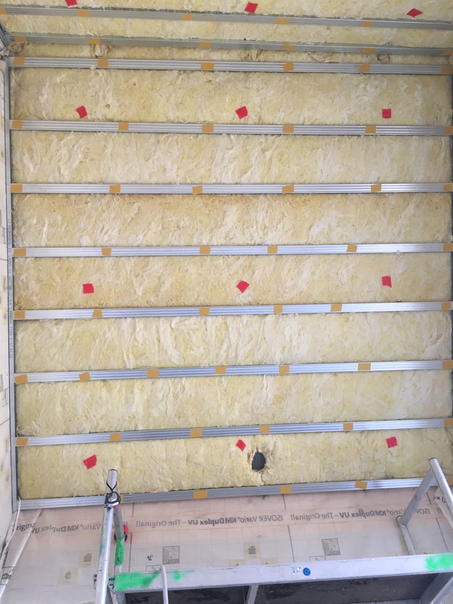 Poddasze w trakcie ocieplania wełną mineralną, widoczne metalowe profile mocowane do sufitu z przyklejonymi żółtymi taśmami, czerwone znaczniki i fragment drabiny na dole zdjęcia.