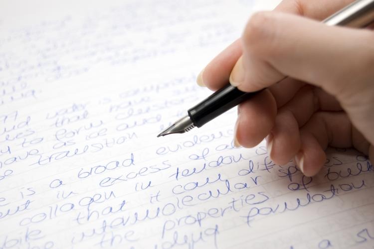 Ręka pisząca niebieskim atramentem piórem wiecznym na papierze w linie, fragment tekstu widoczny.