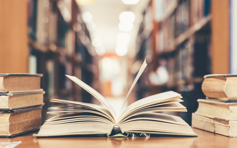 Otwarta książka na drewnianym stole w bibliotece, z rzędami książek w tle, uchwycona w ciepłym oświetleniu.