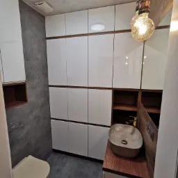 Nowoczesna łazienka z szarymi płytkami imitującymi beton, białymi szafkami z drewnianymi akcentami, umywalką nablatową w odcieniach beżu oraz dekoracyjnymi żarówkami na sznurach.