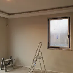 Pokój po malowaniu ścian na szaro, zabezpieczone okna folią, widoczne dwie drabiny aluminiowe.