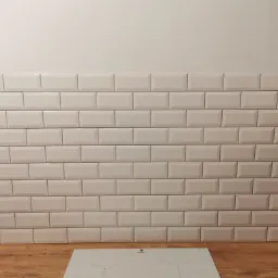 Ściana wyłożona białymi, prostokątnymi płytkami typu metro, z widoczną płytą indukcyjną Electrolux na drewnianym blacie, w tle fragment szafki i wiadro z zaprawą.