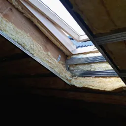 Ocieplenie poddasza wełną mineralną, widoczne metalowe profile i okno dachowe, częściowo wykończone drewnem, z widokiem na dachówkę na zewnątrz.