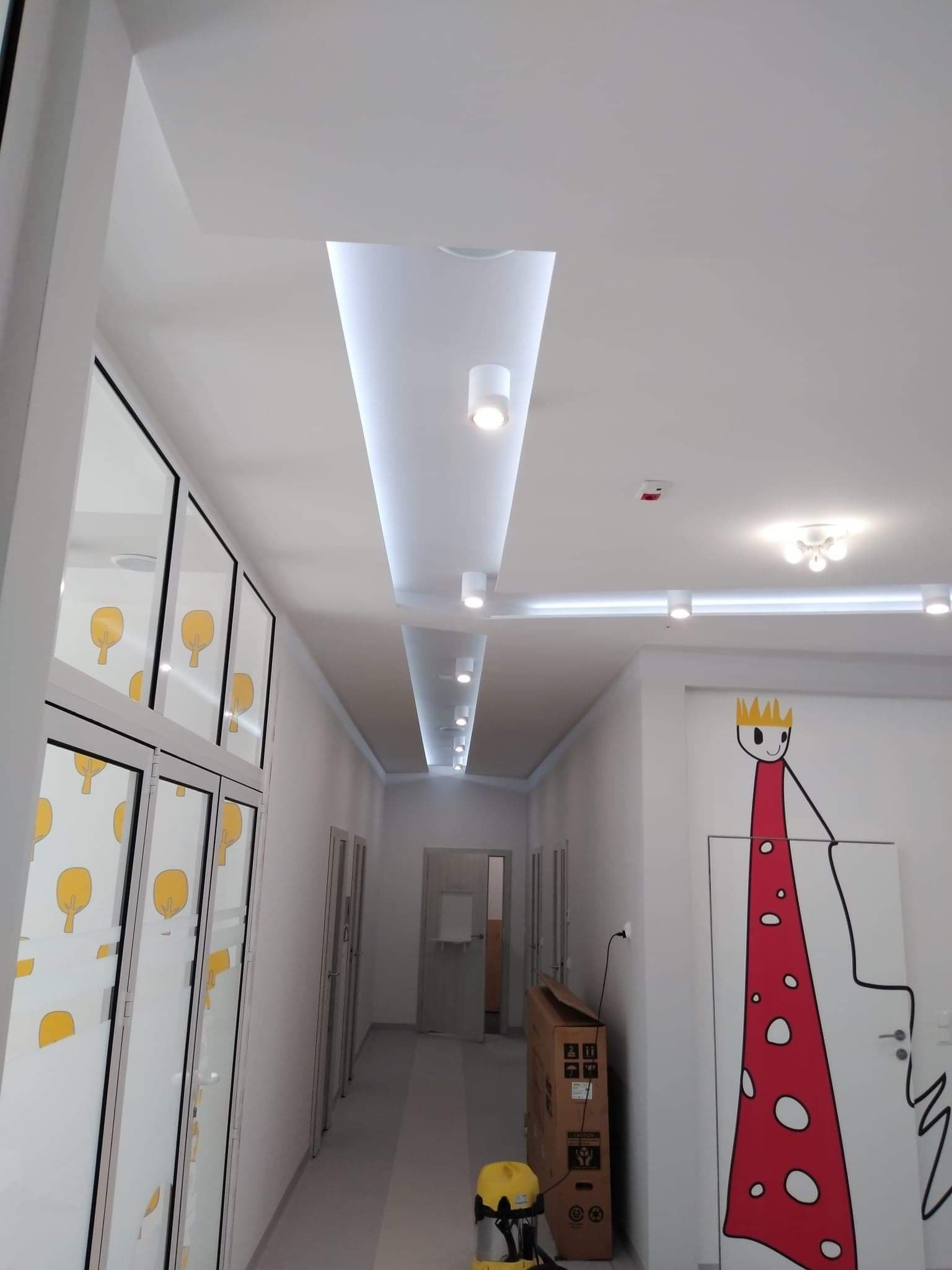 Korytarz w jasnych barwach z podwieszanym sufitem z oświetleniem punktowym i liniowym LED, szklane drzwi z motywem drzewek i malunek na drzwiach przedstawiający postać w koronie, kartonowe pudło...