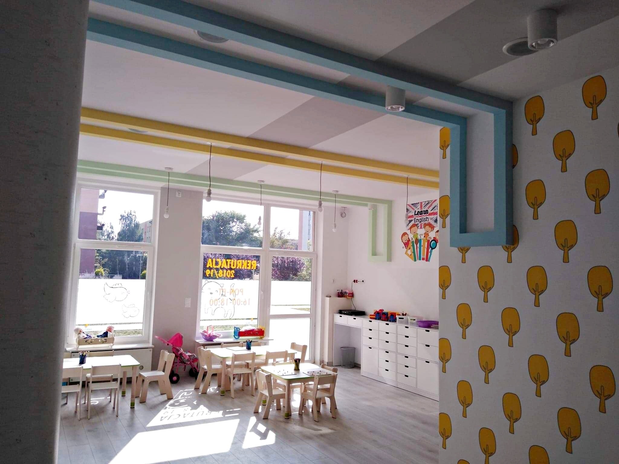 Jasne wnętrze sali przedszkolnej z drewnianymi meblami, białymi szafkami i ścianą z wzorem złotych drzewek; niebiesko-żółto-zielone akcenty kolorystyczne na suficie i ścianach.