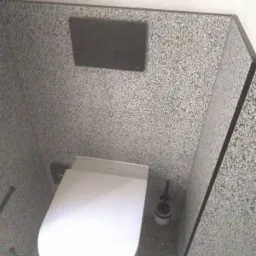 Wisząca toaleta z białej ceramiki, zamontowana na ścianie wyłożonej mozaiką w odcieniach szarości, z czarnym przyciskiem spłukującym nad nią.