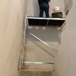 Pracownik na rusztowaniu wyrównuje ścianę gładzią w wąskim korytarzu na schodach, widoczne ślady po wcześniejszych pracach i niedoskonałości ścian.