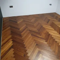 Ukończona podłoga z parkietu ułożonego we wzór jodełki francuskiej, widoczne gniazdka elektryczne na białej ścianie i czarny fragment ściany po lewej stronie.