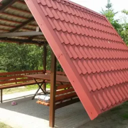 Altana z brązową konstrukcją i czerwoną dachówką, otoczona zielenią, z drewnianymi ławkami i stołem wewnątrz.