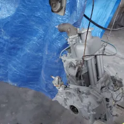 Silnik spalinowy po piaskowaniu i malowaniu na szaro, wiszący na linie na tle niebieskiej plandeki, widoczne ślady farby na podłodze i ścierka.