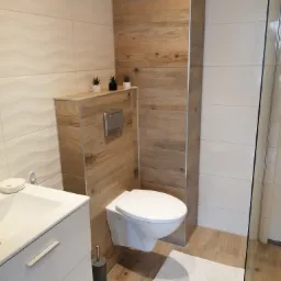 Nowoczesna łazienka z podwieszaną toaletą obudowaną drewnopodobną płytą, białą umywalką z szafką i dekoracyjnymi roślinami w doniczkach.