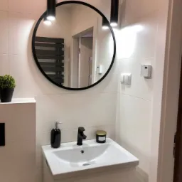 Nowoczesna łazienka z białymi płytkami, okrągłym lustrem w czarnej ramie, czarną armaturą i cylindrycznymi lampami wiszącymi.