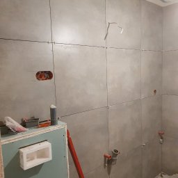 Dawid Cierlik - Łazienka w trakcie remontu: szare, matowe płytki ułożone na ścianach z krzyżykami dystansowymi, widoczne instalacje hydrauliczne i elektryczne, zabudowa z płyty karton gips z wnęką, poziomica...