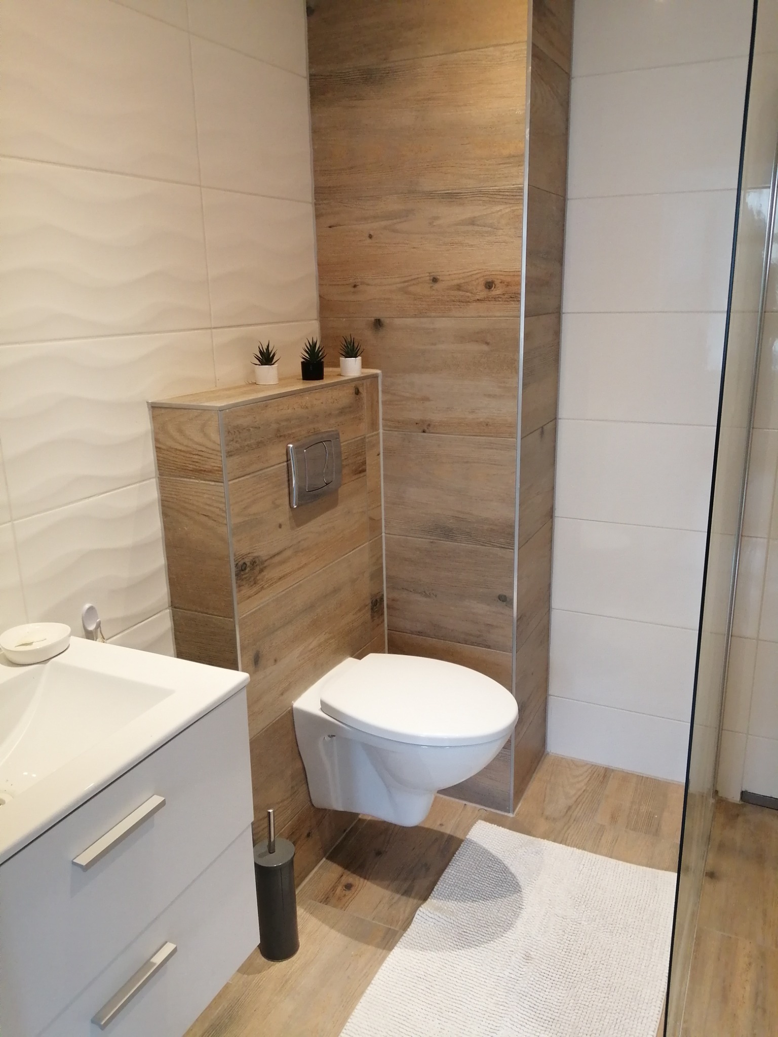 Nowoczesna łazienka z podwieszaną toaletą obudowaną drewnopodobną płytą, białą umywalką z szafką i dekoracyjnymi roślinami w doniczkach.