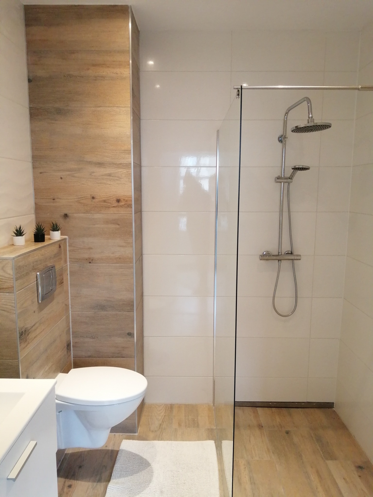 Nowoczesna łazienka z kabiną prysznicową typu walk-in, wykończona płytkami imitującymi drewno na podłodze i częściowo na ścianie, oraz białymi płytkami na pozostałych ścianach, z minimalistycznym...