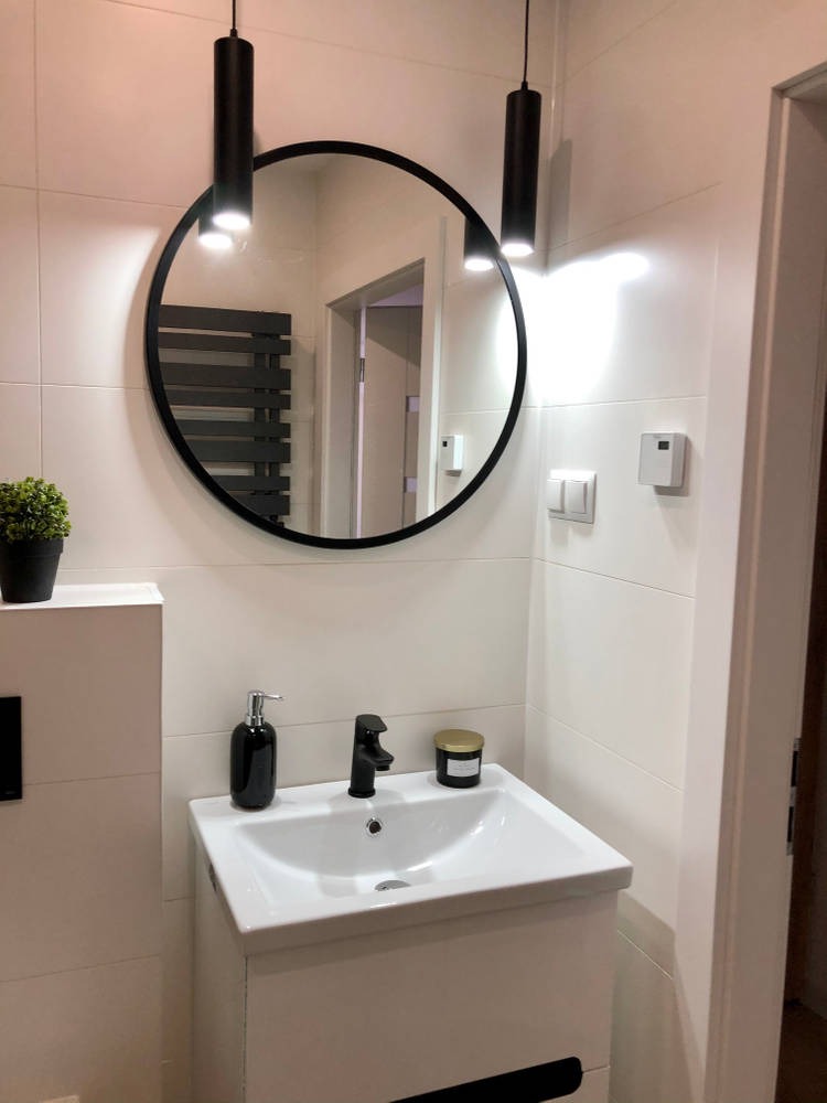 Nowoczesna łazienka z białymi płytkami, okrągłym lustrem w czarnej ramie, czarną armaturą i cylindrycznymi lampami wiszącymi.