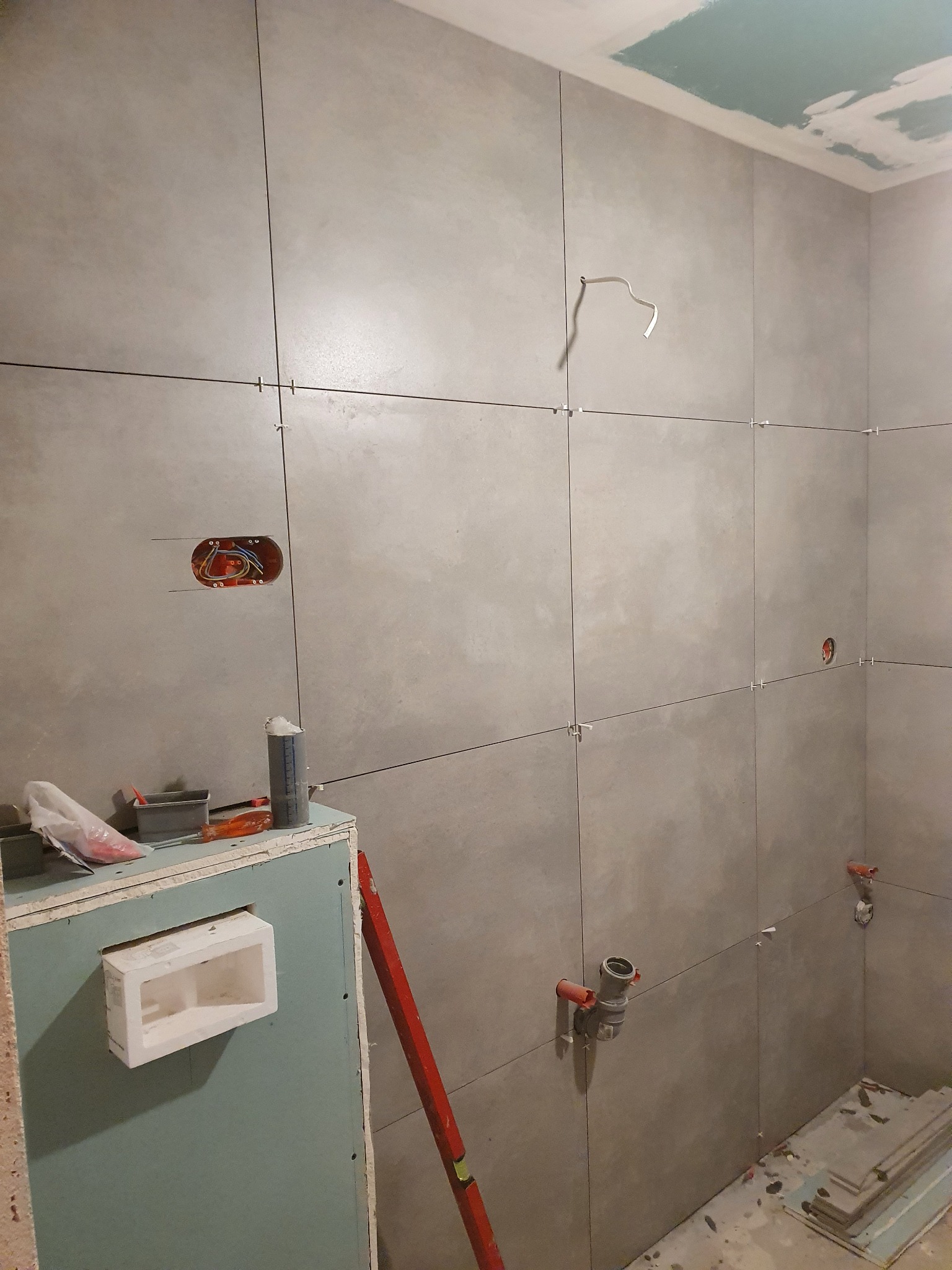 Łazienka w trakcie remontu: szare, matowe płytki ułożone na ścianach z krzyżykami dystansowymi, widoczne instalacje hydrauliczne i elektryczne, zabudowa z płyty karton gips z wnęką, poziomica...