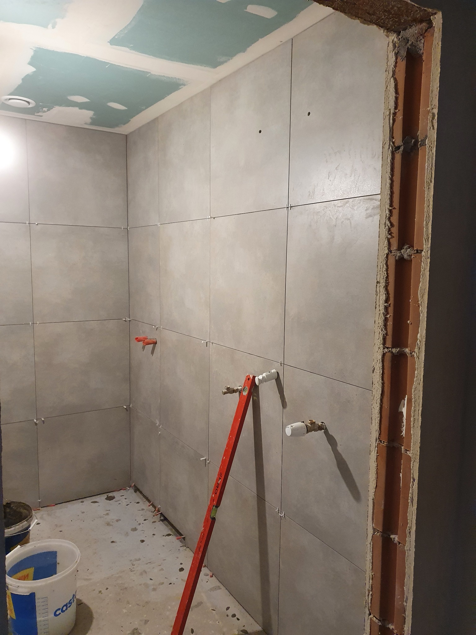 Świeżo wykafelkowana ściana w łazience z widocznymi wystającymi rurami, poziomica oparta o ścianę, widoczne plastikowe krzyżyki dystansowe między płytkami i surowy otwór drzwiowy z cegły.