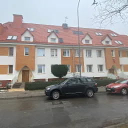 Budynek wielorodzinny po remoncie elewacji z elementami imitującymi drewno, widoczny cokół z płytek klinkierowych, czerwona dachówka i trzy zaparkowane samochody.