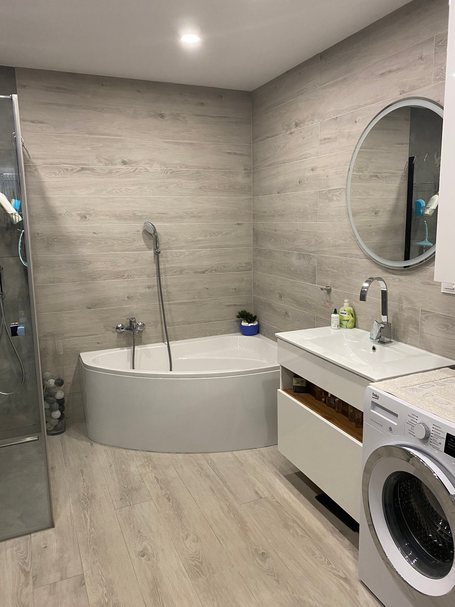 Nowoczesna łazienka z wanną narożną, umywalką na szafce z otwartą półką, okrągłym lustrem z podświetleniem LED i pralką Beko, wykończona płytkami imitującymi drewno.