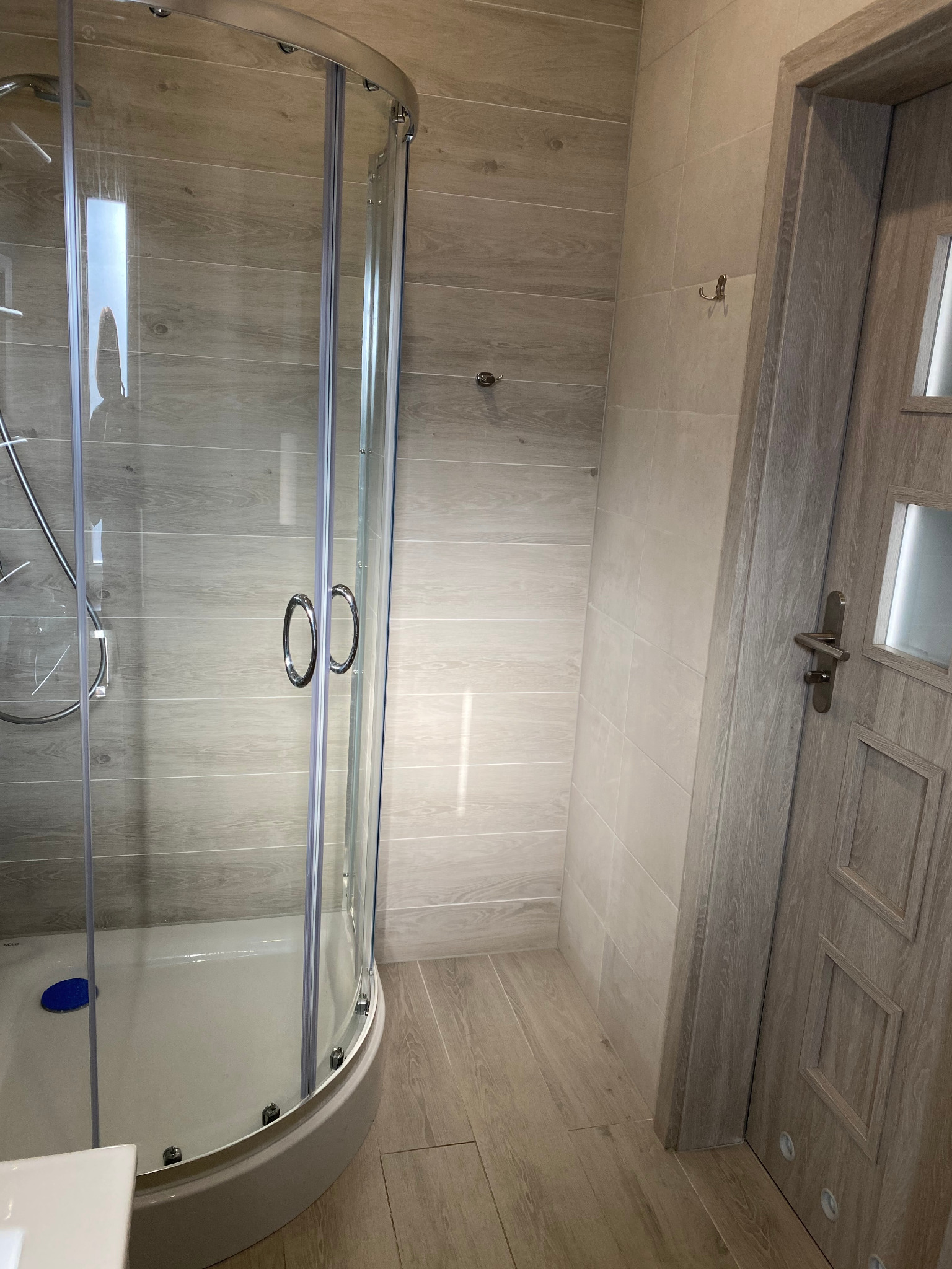 Nowoczesna kabina prysznicowa z zaokrąglonymi drzwiami i chromowanymi uchwytami, wykończona płytkami imitującymi drewno, z widoczną podłogą i drzwiami w odcieniach szarości.