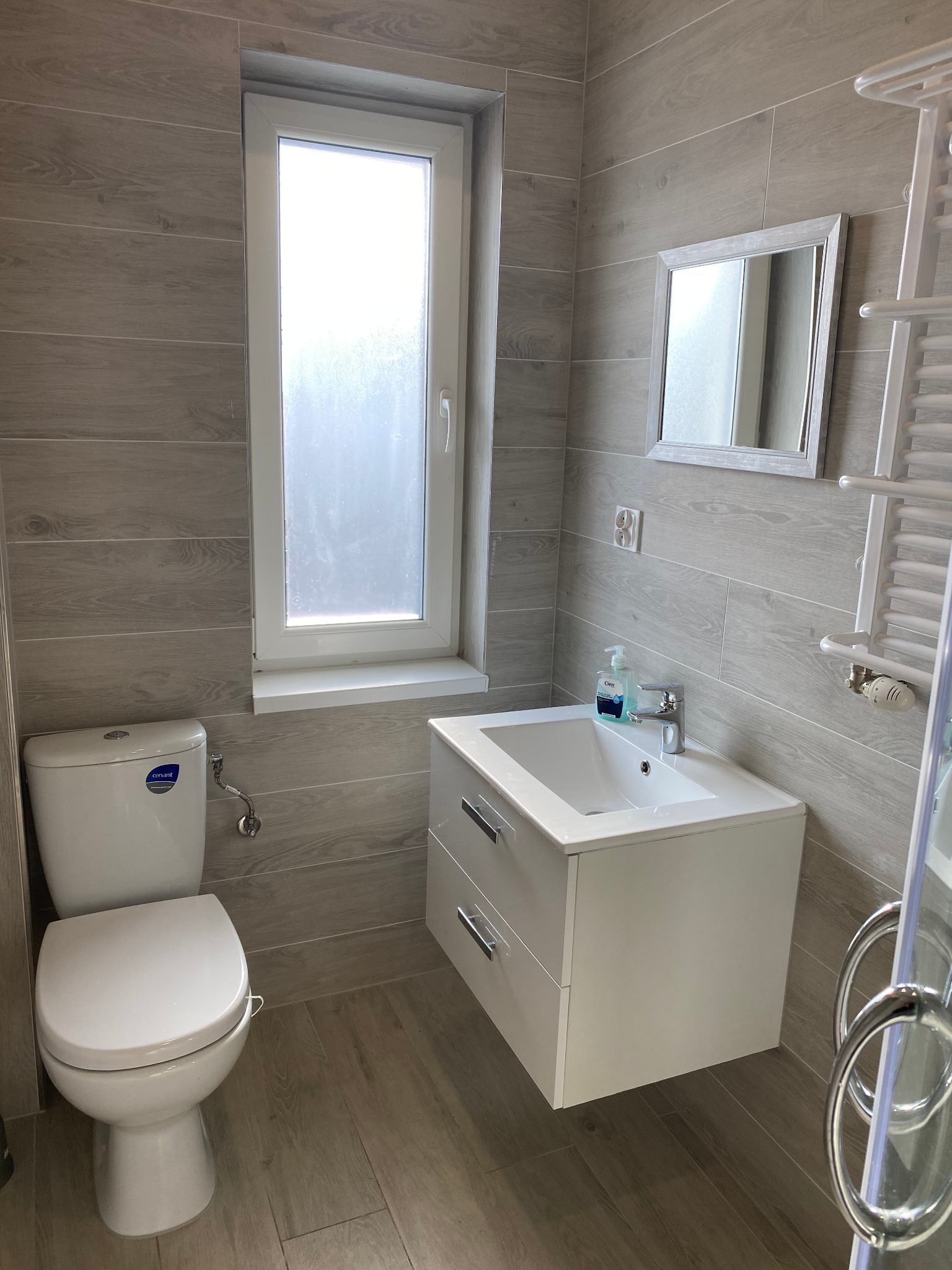 Nowoczesna łazienka z toaletą Cersanit, białą szafką pod umywalkę z dwiema szufladami, lustrem w szarej ramie i grzejnikiem drabinkowym, wykończona płytkami imitującymi drewno.