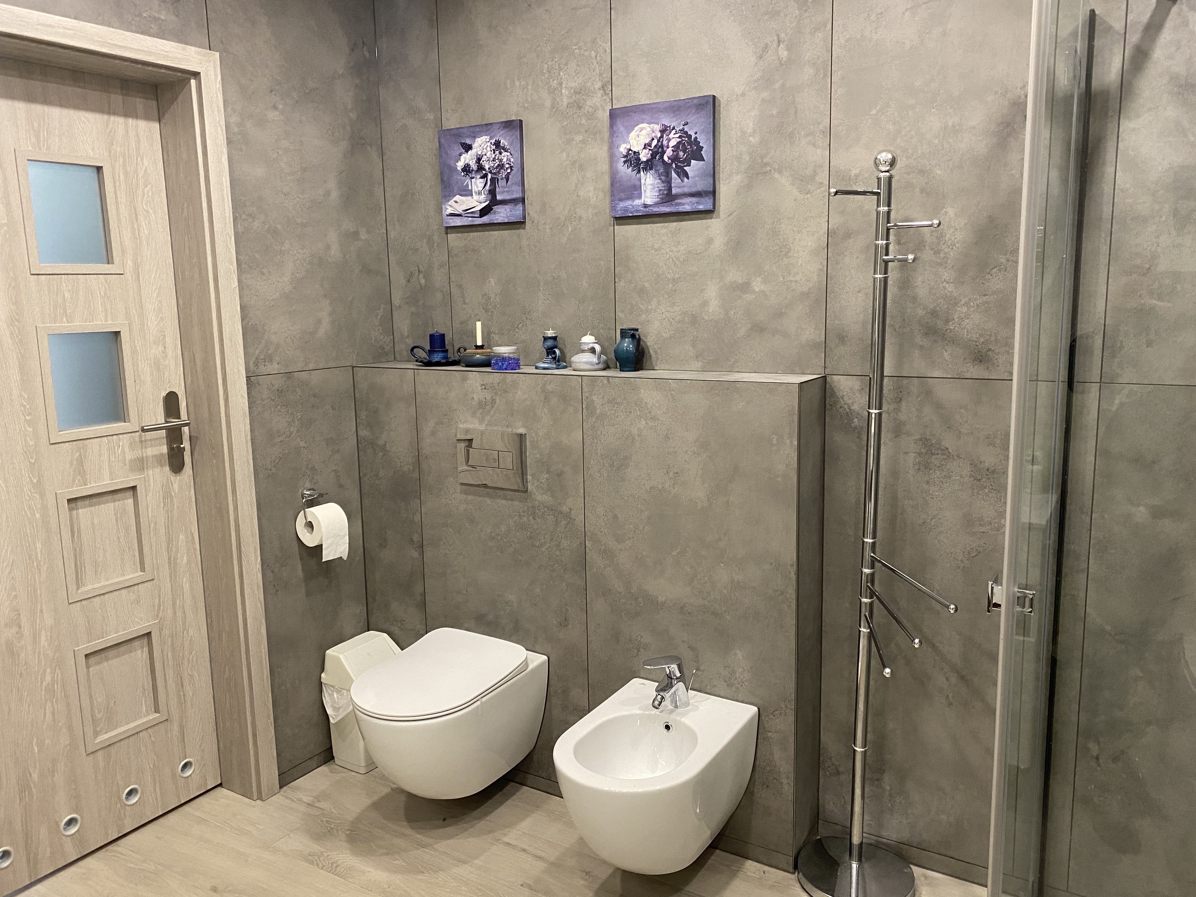 Nowoczesna łazienka z szarą, betonową teksturą ścian, podwieszaną toaletą i bidetem, dekoracyjnymi obrazami kwiatów i chromowanym wieszakiem na ręczniki.