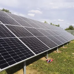 Instalacja paneli słonecznych na metalowych wspornikach na łące, w tle bele siana owinięte folią.