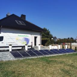 Manufaktura Energii Artur Mrozek - Instalacja paneli słonecznych na gruncie obok domu jednorodzinnego, z widocznym firmowym vanem z logo i danymi kontaktowymi, ustawionym obok dekoracyjnych donic.