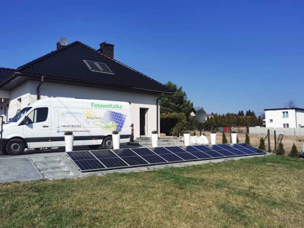 Instalacja paneli słonecznych na gruncie obok domu jednorodzinnego, z widocznym firmowym vanem z logo i danymi kontaktowymi, ustawionym obok dekoracyjnych donic.