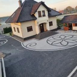 Nowoczesny dom z efektownym podjazdem z kostki brukowej w odcieniach szarości, z dekoracyjnym motywem kwiatowym i spiralnymi wzorami, widok z góry w porannym świetle.