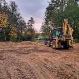 Żółta koparko-ładowarka na placu budowy w Chrzypsku Wielkim, widoczne ślady opon w ziemi i usypane pryzmy ziemi, drzewa w tle pod pochmurnym niebem.