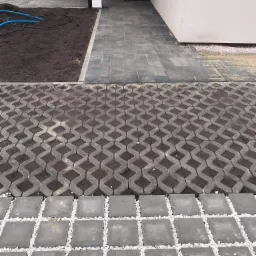 Ułożona nawierzchnia z kostki brukowej w trzech różnych wzorach: prostokątna ścieżka, ażurowa kratka wypełniona ziemią i kwadratowa kostka z białym grysem, prowadząca do wejścia budynku z widoczną...