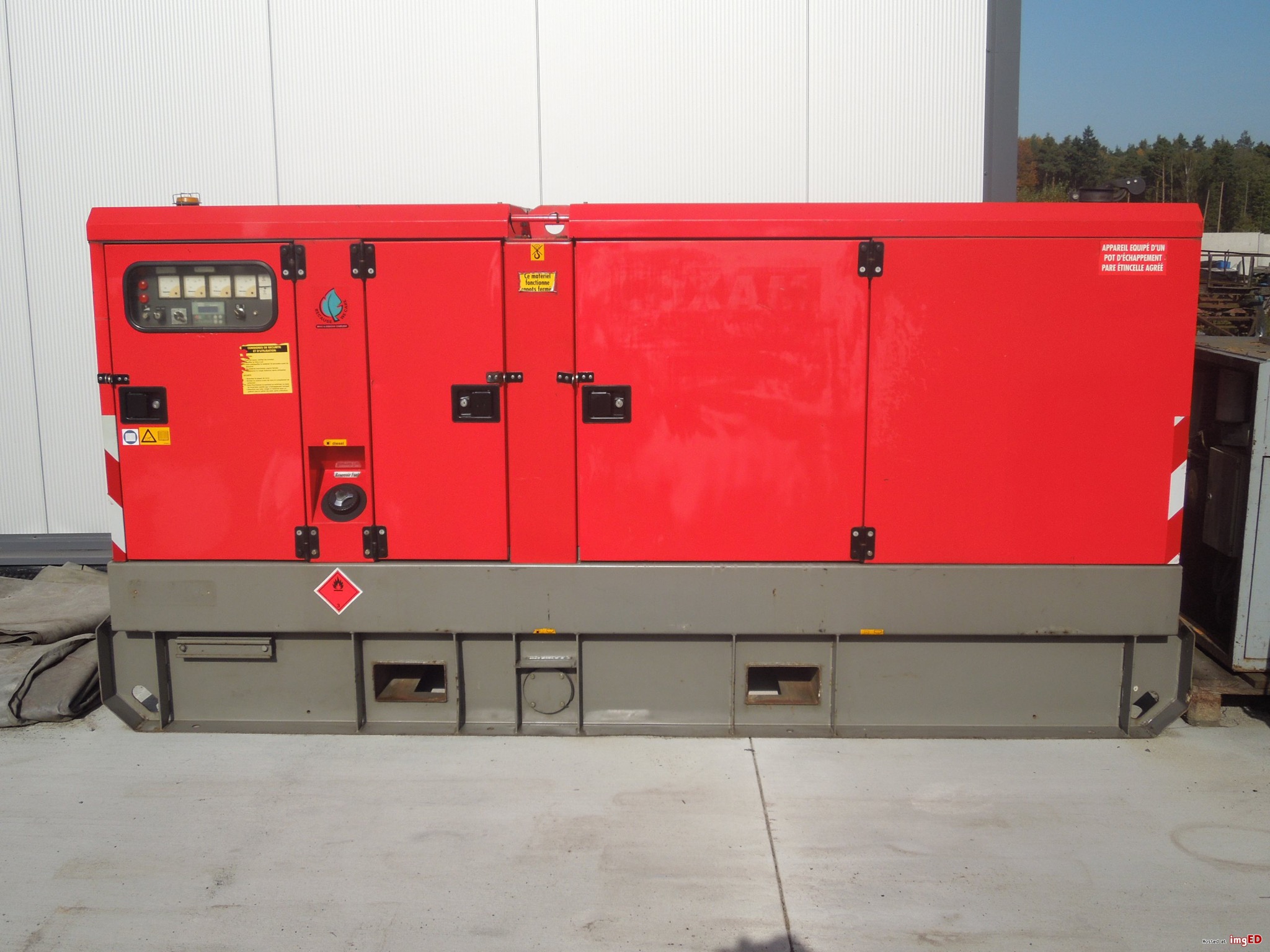 Czerwony, przemysłowy agregat prądotwórczy z widocznymi wskaźnikami, oznaczeniami ostrzegawczymi i zamkniętymi drzwiami, umieszczony na szarym fundamencie.
