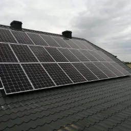 Panele słoneczne na ciemnym, metalowym dachu domu jednorodzinnego, widok z dołu, w tle pochmurne niebo i pole.