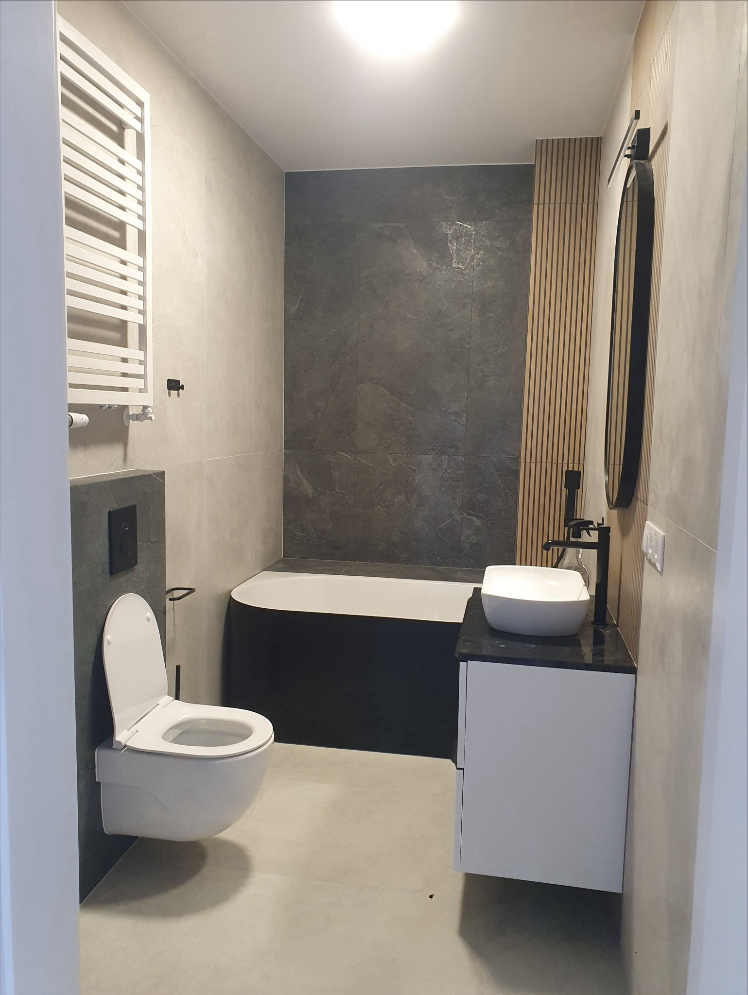 Nowoczesna łazienka w Warszawie: czarna wanna, umywalka na szafce, wisząca toaleta i grzejnik drabinkowy na tle szarych płytek i drewnianych paneli.