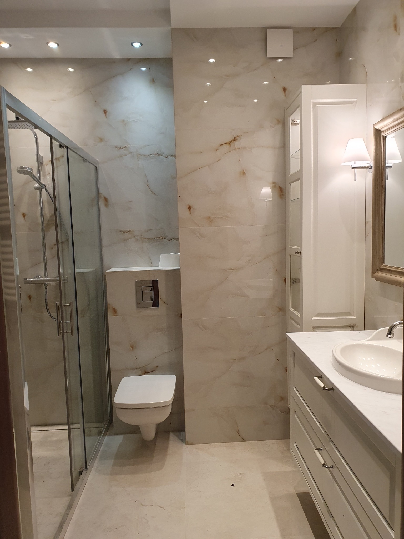 Wykończona łazienka z kabiną prysznicową, toaletą podwieszaną i umywalką z szafką w stylu klasycznym. Płytki imitujące marmur na ścianach i podłodze. Oświetlenie punktowe.