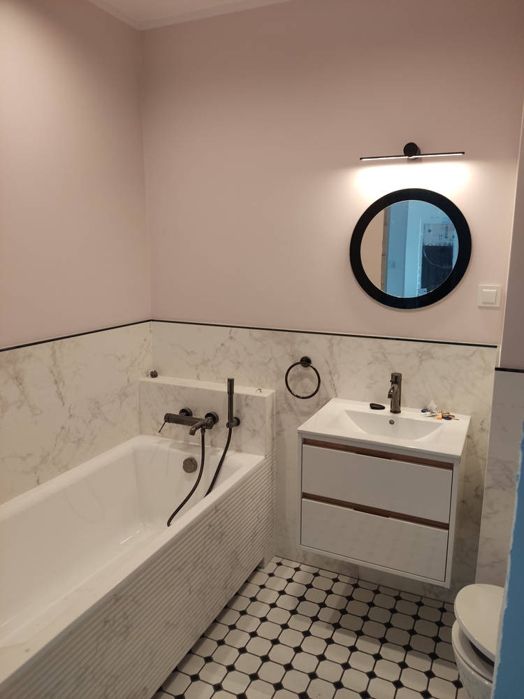 Elegancka łazienka w odcieniach bieli i pudrowego różu. Marmurowe płytki, czarne akcenty armatury i okrągłe lustro z podświetleniem tworzą spójną całość.