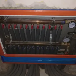 Rozdzielacz ogrzewania podłogowego z zaworami, manometrami i izolowanymi rurami, zamontowany w obudowie z pomarańczowo-niebieskim wykończeniem, z widocznymi szarymi rurami instalacji. Ściana w tle...